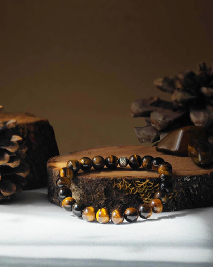 Bracelet en Œil de Tigre naturel posé sur une rondelle de bois avec une pomme de pin en arrière-plan, entouré d’une pierre polie d’Œil de Tigre.