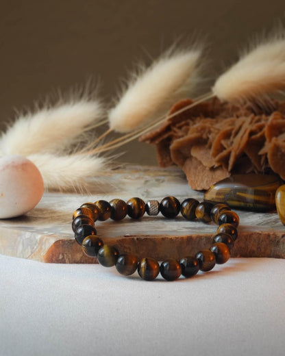 Bracelet en Œil de Tigre naturel posé sur une roche lisse et plate avec des épis blancs en arrière-plan, entouré d’une pierre polie d’Œil de Tigre.